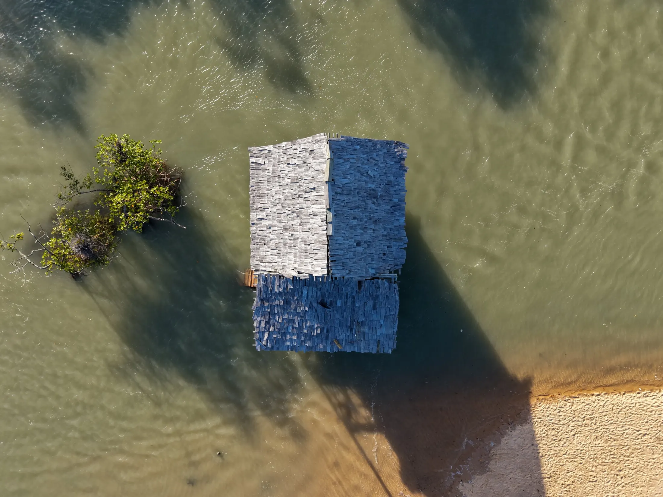 Vista aérea de uma casinha de madeira sobre a água, ao lado de um manguezal, perto da areia