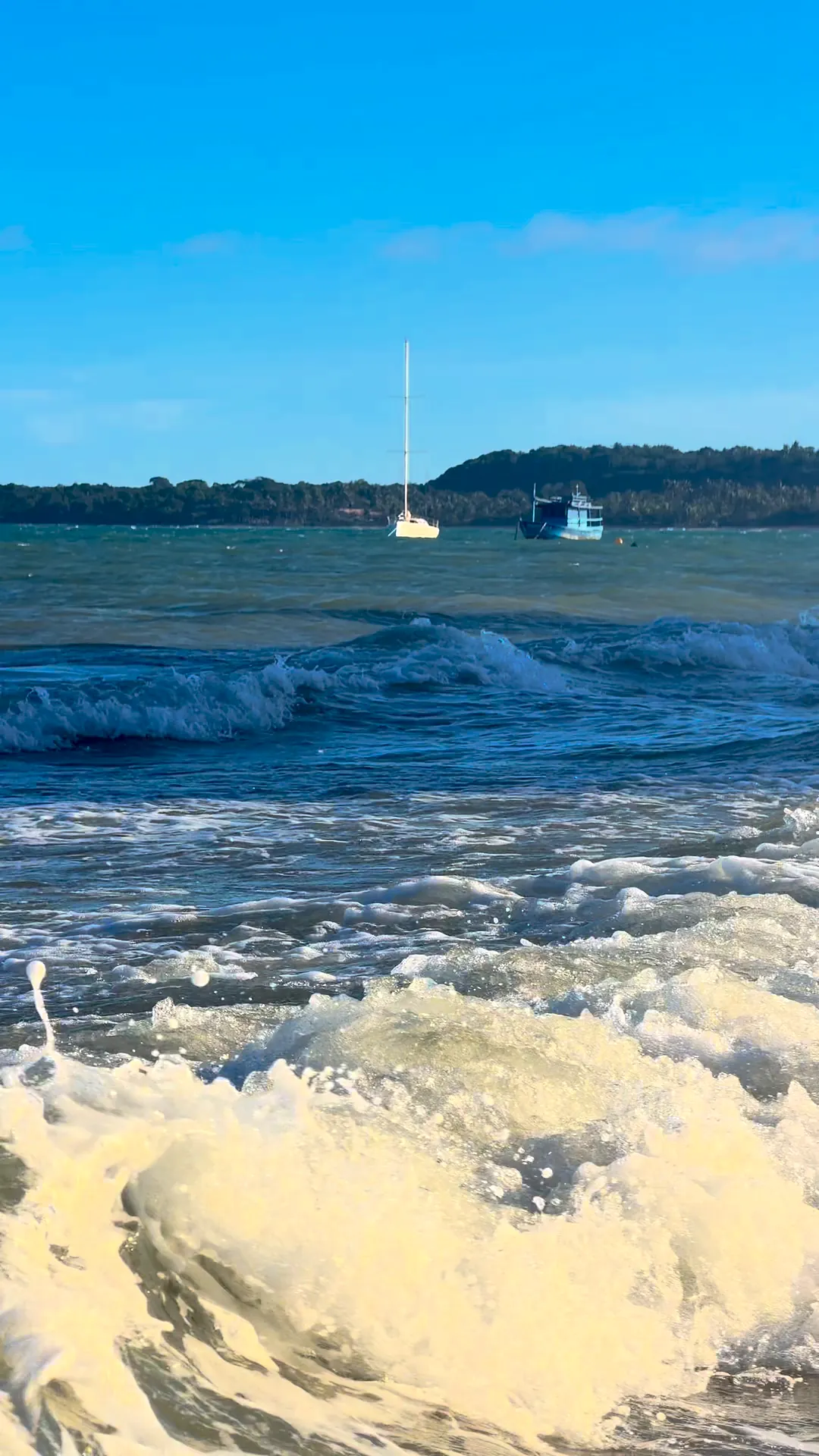 Ondas quebrando na beira do mar, com um veleiro ancorado ao fundo
